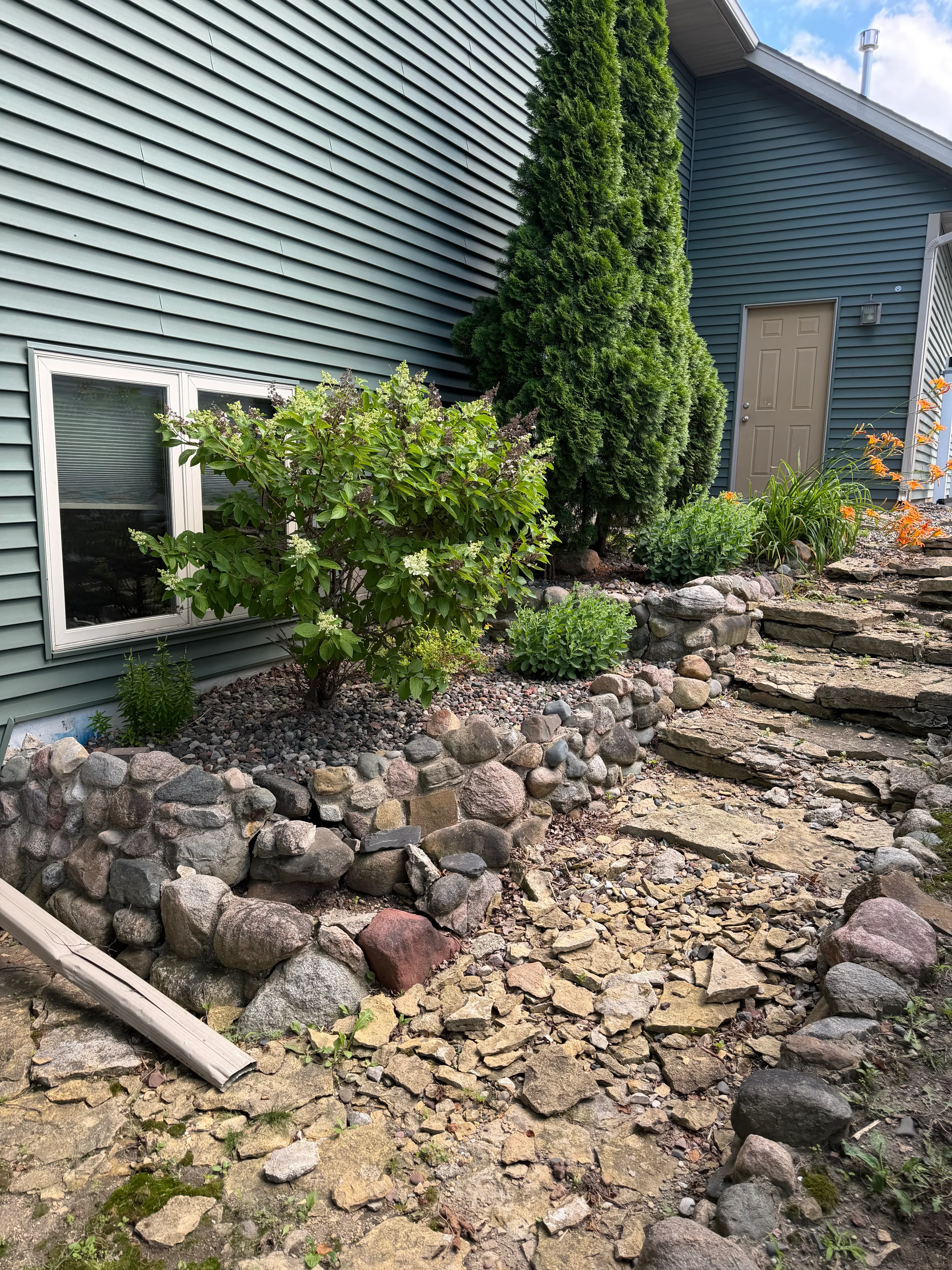 Stacked natural stone retaining wall with green shrubs and ornamental plantings beside metal building